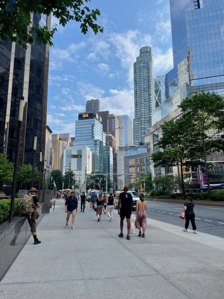 Pedestrians walk along a sidewalk lined with skyscrapers in New York City. The reflective glass facades of modern high-rises tower over the street, while people casually stroll under a clear blue sky with a few clouds.