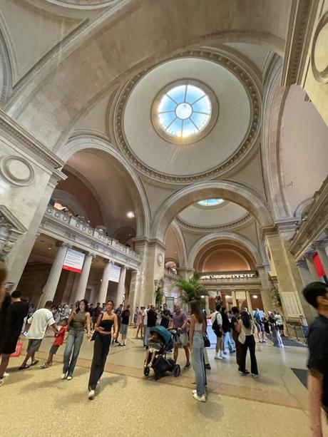 Inside the Metropolitan Museum of Art, visitors walk through the grand hall beneath towering arches and a large central dome skylight. The spacious interior features classical architectural elements, with people exploring the exhibits and admiring the museum's grandeur.