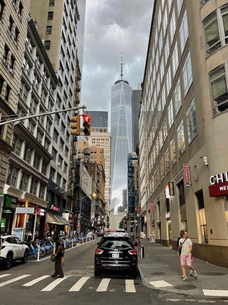 A street-level view of One World Trade Center framed by surrounding buildings, as cars and pedestrians navigate the busy downtown Manhattan street.