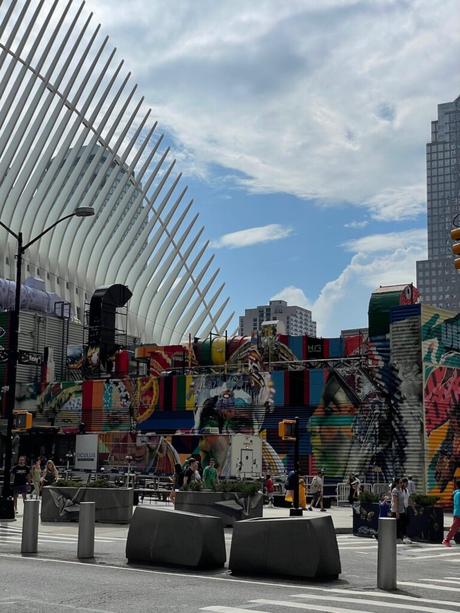 A vibrant mural with colorful patterns and abstract figures near the Oculus transportation hub in New York City, with the iconic white ribs of the Oculus structure in the background.