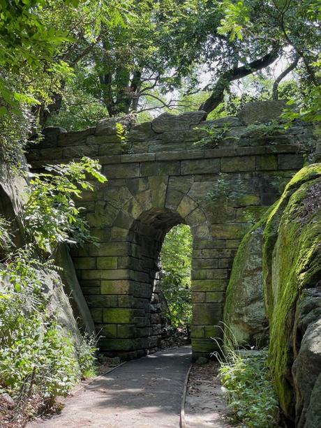 A stone archway nestled within the greenery of Central Park, New York City. The pathway leading under the arch is surrounded by moss-covered rocks and shaded by dense trees.