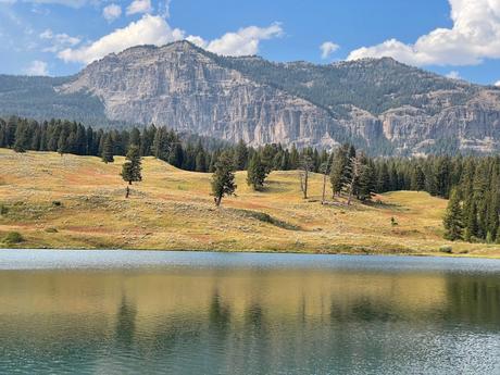 A calm lake reflecting grassy hills and scattered trees, with a tall rocky mountain ridge in the background.