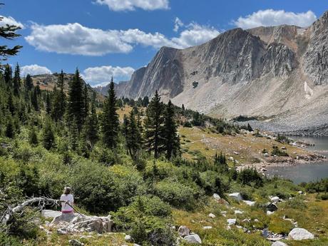 A hiker sitting on a rock, admiring the view of dramatic mountain cliffs and a serene alpine lake, surrounded by lush green forest in the Snowy Range near Centennial, Wyoming.