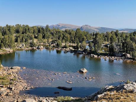 A serene lake surrounded by trees and rocky shores, with distant mountains under a clear blue sky.