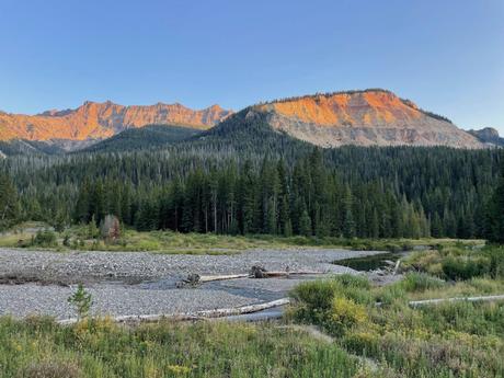 An orange-tinged mountain glowing in the sunset, with a forest of tall evergreens below and a river flowing through a rocky landscape.