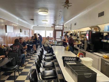 Inside view of a retro diner in Laramie, Wyoming, with checkered floors, counter seating, and customers eating at tables. The staff are visible behind the counter serving food.