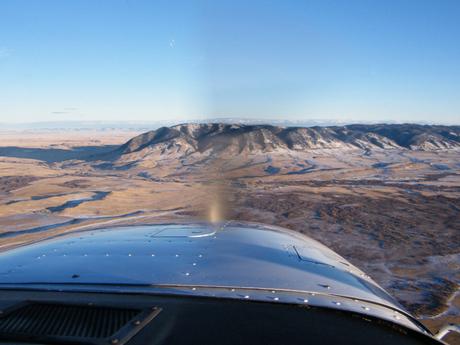 An aerial view from a single engine propellor plane showing wide, open plains leading to a snow-covered mountain range under a bright blue sky.