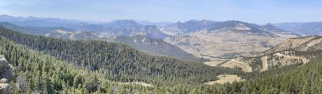 A panoramic view of a winding road through a forested valley with distant mountain ranges under a light blue sky.