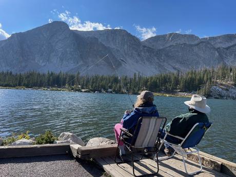 Two people, sitting in folding chairs on a wooden deck, fishing in a calm mountain lake surrounded by towering rocky cliffs and pine trees.