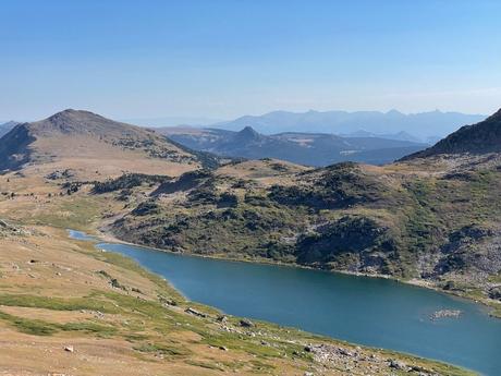 A serene landscape of a lake surrounded by grassy plains and rolling hills, with distant mountain ranges under a bright, clear sky.