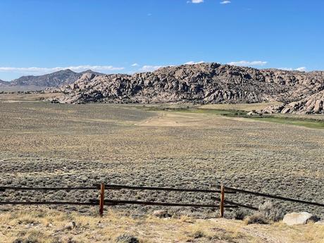 A wide, rocky expanse of grassland with a rustic wooden fence and jagged rock formations in the background under a bright sky.