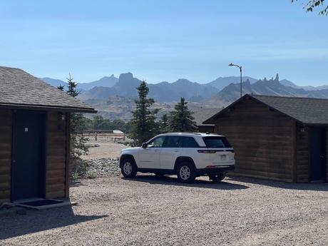 A white SUV parked between two wooden cabins with a backdrop of rugged, mountainous terrain under a clear blue sky.