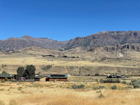 A dry, open landscape with mountains in the distance and a few scattered homes and structures in the foreground, under a bright blue sky.