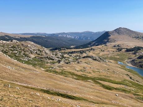 A wide view of a rolling hillside with a lake in the distance, rocky terrain, and grassy slopes, with mountain ranges fading into the horizon.