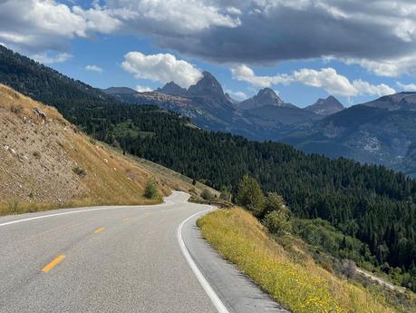 A winding highway cuts through the foothills, leading toward the Tetons under a partly cloudy sky, with forested hills in the foreground.