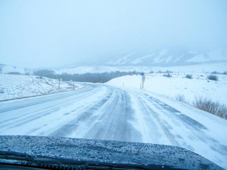 A snow-covered road in the mountains, the view partially obscured by snow, with snow-covered hills in the background.