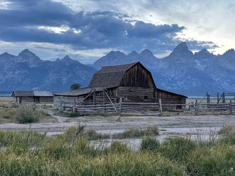 An old wooden barn sits in an open field at Mormon Row, with the jagged peaks of the Grand Tetons rising dramatically behind it.