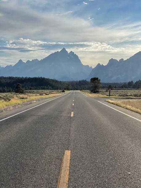 A straight road leading toward the distant Grand Teton mountains under a partly cloudy sky, with trees on either side of the road.