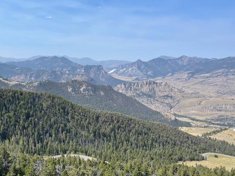 A mountain range viewed from above, with forested hills in the foreground and expansive peaks in the background under a light blue sky.