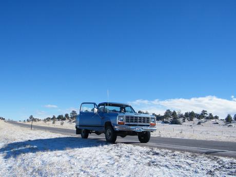 A blue pickup truck with its door open, parked on a snow-dusted roadside under a clear blue sky, surrounded by sparse trees.