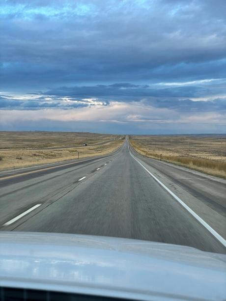 A long, straight highway through Wyoming disappearing into the horizon under a cloudy sky, with vast open plains on either side of the road.