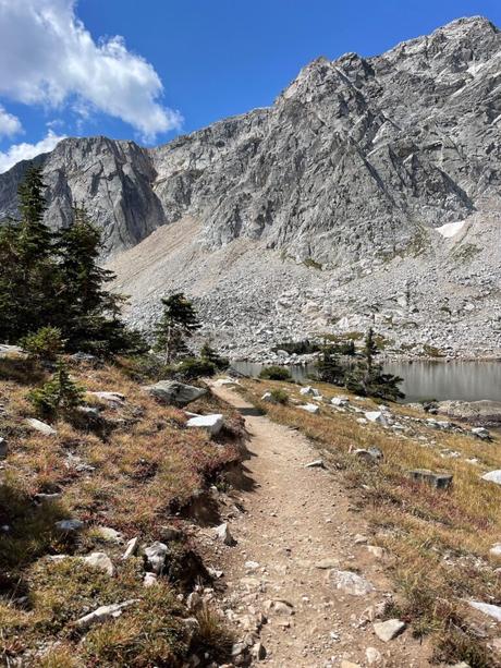 A rocky dirt trail winding through a mountain landscape in the Snowy Range in southern Wyoming, with steep, jagged cliffs towering in the background under a blue sky.