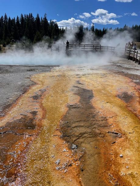 Steaming hot springs with vibrant orange and yellow mineral deposits on the ground, with people observing from a wooden walkway.