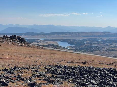 A panoramic view of a wide, open plain with a distant lake, rock formations, and rolling hills stretching into the distance.