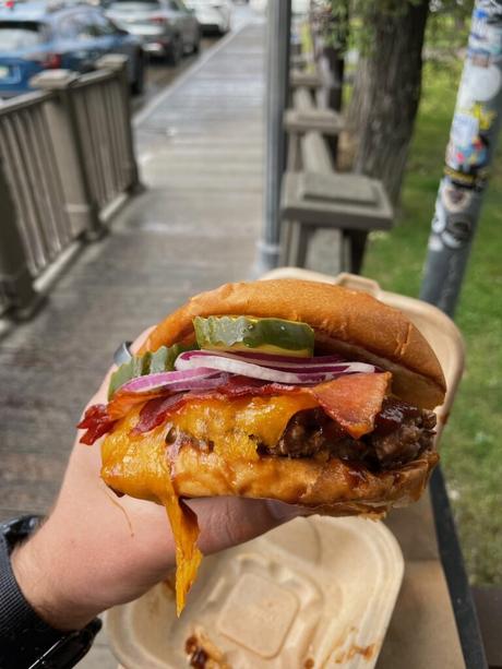 Close-up of a juicy bison burger held in one hand, with melted cheese, crispy bacon, pickles, and onions.