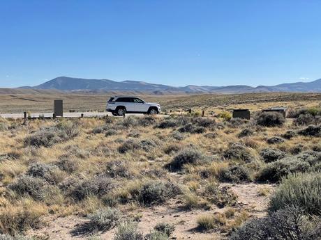 A white SUV parked in a vast open landscape with dry grasses and distant mountains under a clear blue sky.