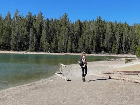 A woman walking along a sandy lakeshore, surrounded by a pine forest and a calm, turquoise lake, wearing a backpack and looking back toward the camera.