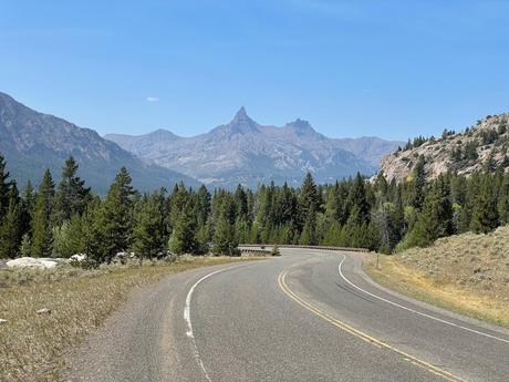 A winding road leading into the mountains, surrounded by trees with jagged peaks in the background.