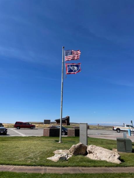 Two flags, the U.S. and Wyoming state flags, waving on a flagpole at a rest area under a clear, bright blue sky.