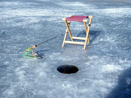 A fishing setup on a frozen lake, with an ice-fishing hole, a rod, and a small wooden stool placed on the ice.