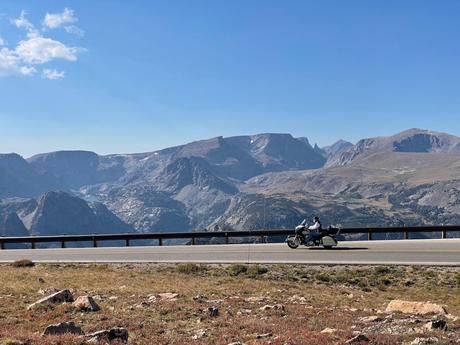 A motorcyclist rides along a scenic mountain road with towering rocky peaks in the background, showcasing a clear, crisp day.