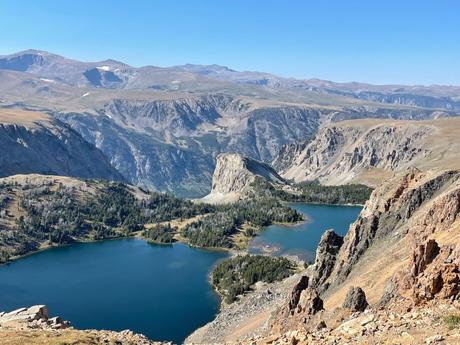 A breathtaking view of a mountain range from the Beartooth Highway with two large lakes nestled among the peaks and valleys, surrounded by rugged cliffs.