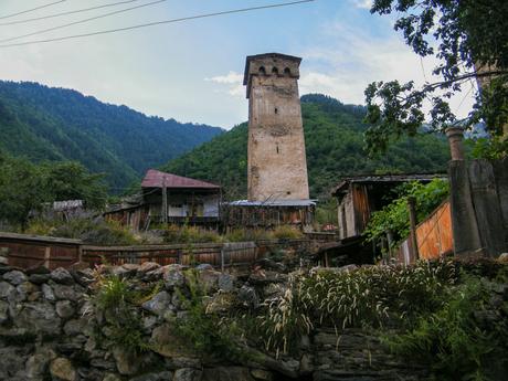 A stone watchtower surrounded by wooden houses in a small Georgian village nestled in a forested mountain region.