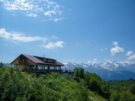 A wooden lodge stands perched on a hillside, its large windows offering a panoramic view of the distant snow-covered mountains under a bright blue sky.