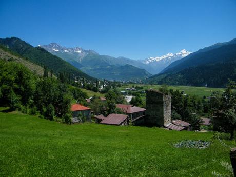 A picturesque valley unfolds beneath a vibrant blue sky, with snow-capped peaks in the distance and a scattering of homes nestled among the green hills.