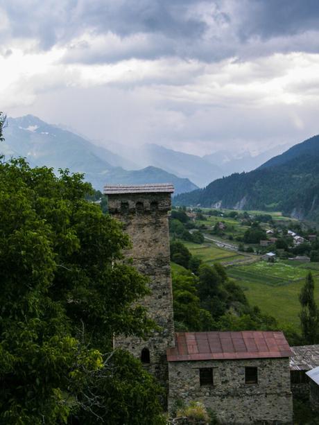 A stone watchtower rising above trees and houses in Svaneti, with green mountains and a cloudy sky in the background.