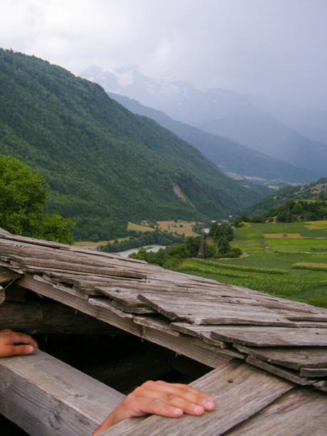 A mountain valley view from the wooden roof of an old Svan tower in Upper Svaneti, with hands gripping the edge of the roof and forested hills below, leading towards distant snow-capped peaks.