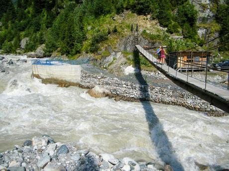 A narrow suspension bridge hangs precariously over a rushing, frothy river, with two hikers crossing, their figures small against the power of the water.