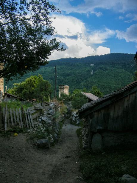 A narrow, dirt path winds through a quiet village, with stone houses and a watchtower nestled among verdant hills, the sky hinting at the end of the day.