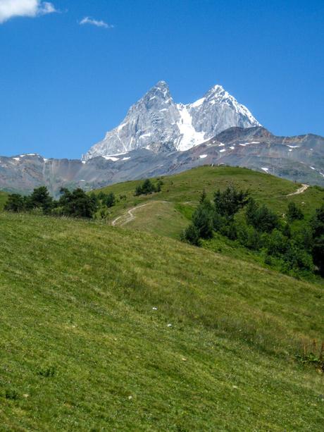 A striking mountain with jagged, twin summits rises dramatically against the clear sky, framed by rolling green pastures and a solitary path leading into the distance.