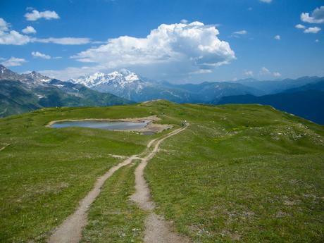 A dirt path leading towards a small mountain lake, with expansive green hills and distant snow-covered mountains in Georgia under a blue sky with clouds.