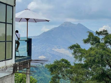 A man sitting on a glass-walled balcony with an umbrella, overlooking a volcanic mountain in the distance.