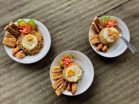 Three plates of traditional Indonesian food, featuring fried noodles, rice, fried egg, tofu, and tempeh, garnished with cucumber and tomato slices.