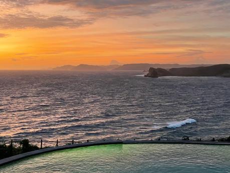 A stunning sunset over the ocean, casting vibrant orange and pink hues across the sky. The water reflects the colors of the sky, with gentle waves rolling toward the shore and there's an infinity pool in the foreground.