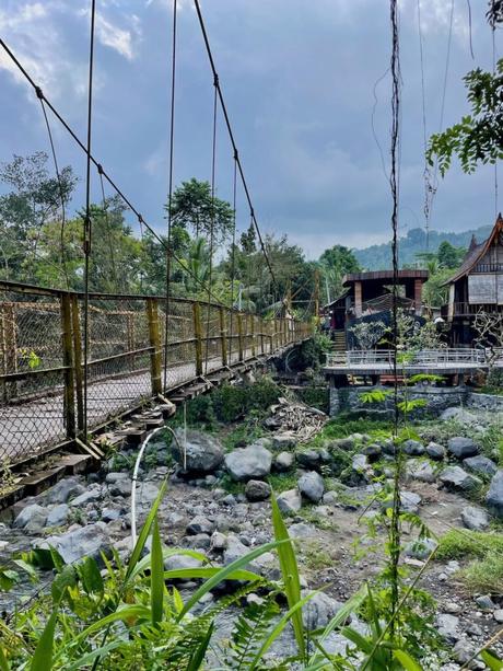 A suspension bridge crossing a rocky stream, leading to wooden houses, with greenery and mountains in the distance.