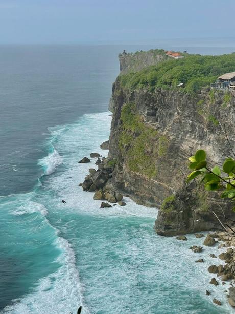 A breathtaking view of the rocky cliffs at Uluwatu, Bali, with turquoise waves crashing against the rocks and the temple visible in the distance.